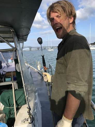 Das Bild zeigt einen Mann, der auf einem Boot steht und eine Angelrute in der Hand hält. Im Hintergrund ist eine Brücke zu sehen und eine Wasserfläche unter einem blauen Himmel mit Wolken.