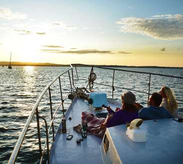 Das Bild zeigt eine heitere Szene mit drei Personen auf einem Boot, die auf das Wasser hinausschauen, während die Sonne in der Ferne untergeht und ein warmes Licht wirft.