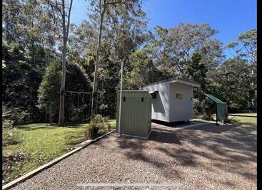 A small white cabin and green shed stand on a gravel patch surrounded by lush trees, grassy lawn, and blue sky, creating a peaceful nature retreat scene.
