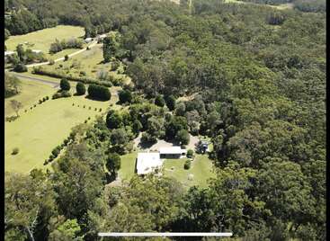 This aerial image shows a house surrounded by lush greenery, dense forest, manicured lawns, a winding road, and scattered trees within a peaceful, rural landscape.