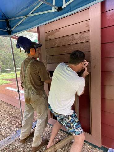 Two men are working on installing or repairing a wooden wall panel outdoors. One wears earmuffs and overalls, while the other kneels, holding the board steady.