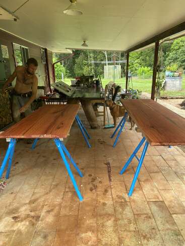 A shirtless man works under a covered patio with wooden planks on blue sawhorses. A brown dog stands nearby. Plants and outdoor furniture surround them.