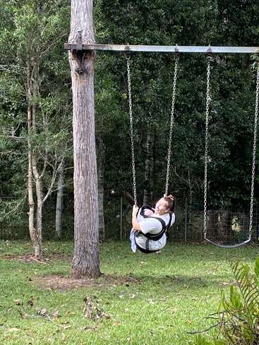 A woman sits on a swing in a grassy yard surrounded by trees, smiling and enjoying herself. The metal swing set is attached to a tree.