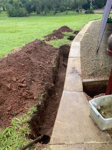 A freshly dug trench runs alongside a concrete path in a garden. Soil mounds border the trench, with grass and trees visible in the background.