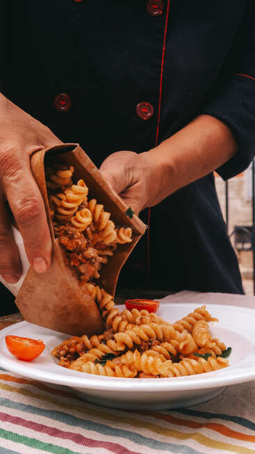Un chef vêtu d'un uniforme sombre verse des fusilli accompagnés de sauce, tirés d'une boîte à emporter, dans une assiette garnie de tranches de tomates, préparant ainsi un délicieux repas prêt à être servi.