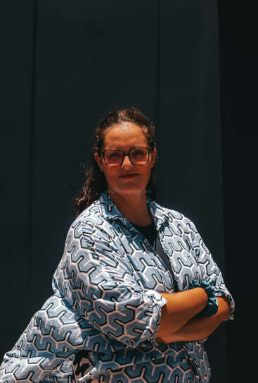 Une femme aux cheveux bouclés, portant des lunettes et une chemise à motifs, se tient debout, l'air assuré, les bras croisés, sur un fond sombre, partiellement baignée de soleil ; elle semble sereine et sûre d'elle.