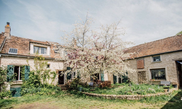 Uma charmosa casa de pedra rústica com persianas verdes, cercada por vegetação. Uma árvore em flor fica no pátio, sob um céu azul claro e brilhante.