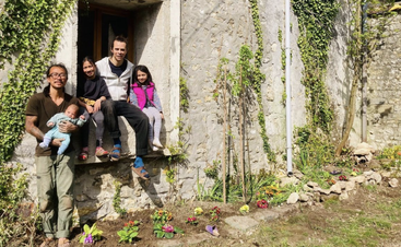 Cinco pessoas - dois adultos e três crianças - estão reunidas do lado de fora de uma construção rústica de pedra coberta de hera. Há flores plantadas embaixo, e todos parecem felizes e relaxados.