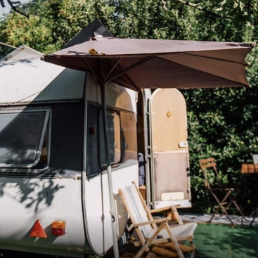 Um campista aconchegante está estacionado ao ar livre com a porta aberta. Duas espreguiçadeiras de madeira estão embaixo de um grande guarda-sol, cercadas por uma folhagem verde exuberante e pela luz do sol.