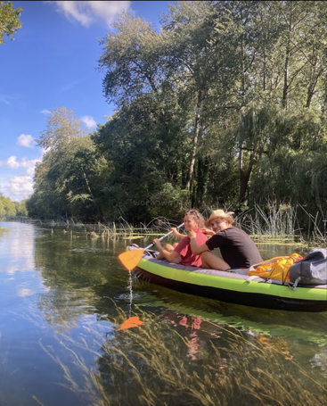 Two people are kayaking on a calm river surrounded by lush green trees under a bright blue sky, enjoying nature and a peaceful outdoor adventure together.
