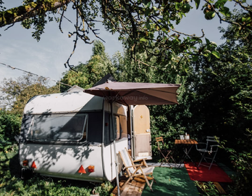 A cozy white caravan sits in a lush green garden, shaded by trees and an umbrella. Nearby, outdoor chairs and a table invite relaxation and leisure.