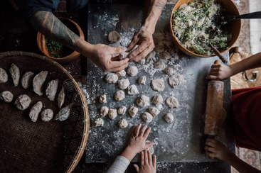 Three people are making dumplings together, working with dough, rolling pin, and vegetable filling. Finished dumplings are placed on a tray, creating a cozy, communal scene.
