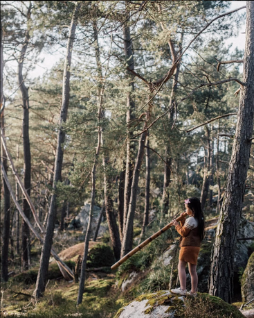 A young girl stands on a mossy rock in a sunlit forest, playing a long wooden wind instrument, surrounded by tall, dense trees and natural light.