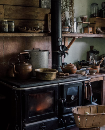 A cozy rustic kitchen with a wood-burning stove, copper kettles, vintage pots, wooden shelves, jars of herbs, utensils, and a basket of apples radiates warmth.