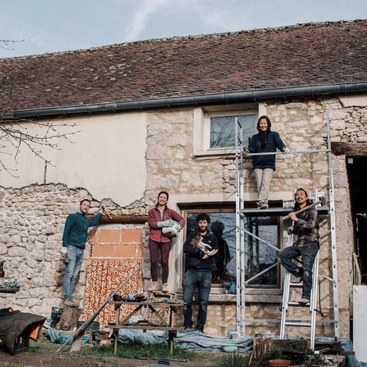 Five people pose in front of an old stone house under renovation. Some stand on scaffolding and a table, smiling, suggesting teamwork and community restoration efforts.