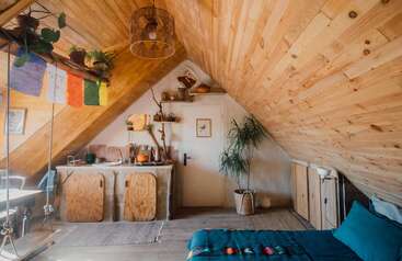 This cozy attic room features wooden walls, a small kitchenette, a bed with teal bedding, potted plants, prayer flags, and a warm, inviting bohemian aesthetic.