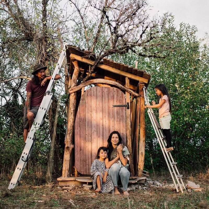 A family of four poses around a rustic wooden structure outdoors. Two children use ladders, while two adults sit and smile amid surrounding trees and nature.
