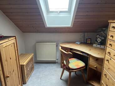 This cozy attic room has wooden furniture, a corner desk, a cushioned chair, a dresser, a skylight window, heating radiator, and slanted wooden ceiling.