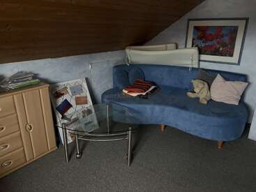 A cozy attic room features a blue curved couch with pillows and a teddy bear, glass coffee table, wooden drawers, framed art, and scattered papers.