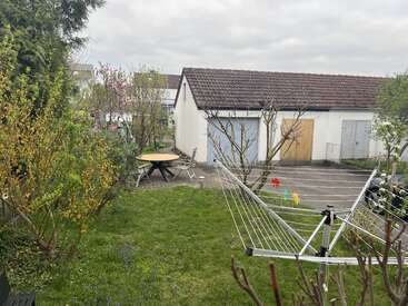 A backyard garden with green grass, trees, and shrubs. There’s a round table, outdoor chairs, a rotary clothesline, and a white garage with closed doors.