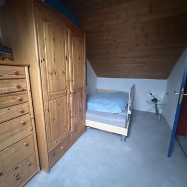 This cozy attic bedroom features wooden furniture, including a wardrobe and chest of drawers, a single bed with blue bedding, slanted wood ceiling, and a minimalist side table.