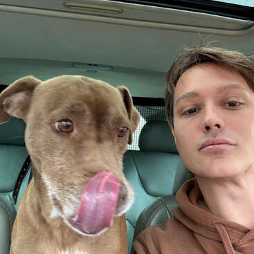 The image shows a selfie of a young man and a brown dog in the back seat of a car, with the dog's tongue out.