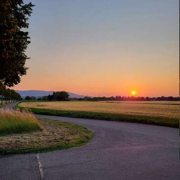 A winding road curves through a peaceful countryside, bordered by trees and fields, while a vibrant orange sunset illuminates the sky and distant hills.