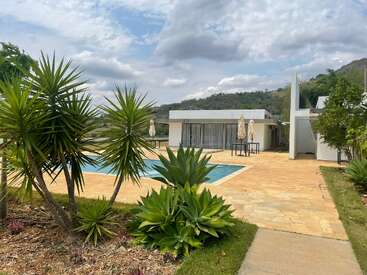 Una moderna casa blanca con grandes puertas de cristal junto a una piscina, rodeada de plantas tropicales y montañas bajo un cielo parcialmente nublado. Relajante entorno al aire libre.