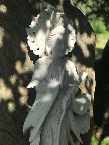 Una serena estatua de piedra blanca de una figura con túnica, posiblemente Kuan Yin, se yergue grácil contra una gran roca, rodeada de luz solar moteada y suaves sombras.