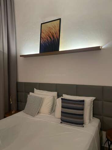 Un dormitorio moderno con una cama bien hecha y almohadas decorativas. Encima, una estantería con luz tenue sostiene una obra de arte enmarcada que representa la naturaleza. Predominan los tonos neutros.