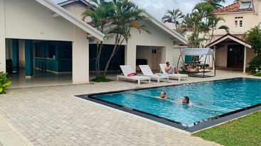La imagen representa una escena serena de una zona de piscina vallada, con una piscina grande y otra más pequeña, rodeadas por una valla, con algunos árboles y una casa al fondo.