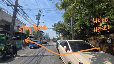 Une rue ensoleillée avec des voitures garées et des arbres bordant les deux côtés. Des notes manuscrites orange et des flèches indiquent les directions : « restez ici » à gauche, « Nous sommes ICI ! » à droite.
