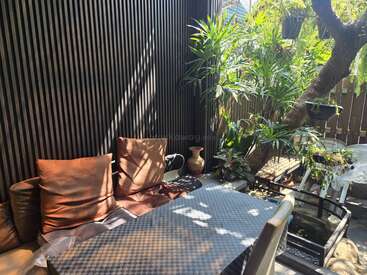 Área externa ensolarada com almofadas marrons, mesa xadrez, plantas verdes exuberantes, parede de ripas de madeira, vasos de cerâmica e um clima tranquilo de jardim. Ambiente relaxante.
