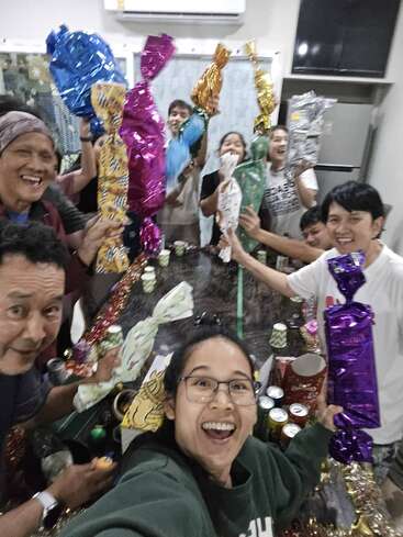 Um grupo de pessoas felizes está reunido em torno de uma mesa, segurando presentes coloridos embrulhados, sorrindo e comemorando, provavelmente curtindo uma festa animada juntos dentro de casa.