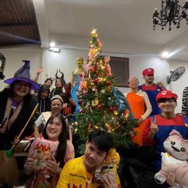 Um grupo animado comemora ao redor de uma árvore de Natal decorada, usando fantasias coloridas, incluindo Mario, Luigi, bruxa e macacões de animais, segurando lanches e brinquedos de pelúcia, sorrindo alegremente.