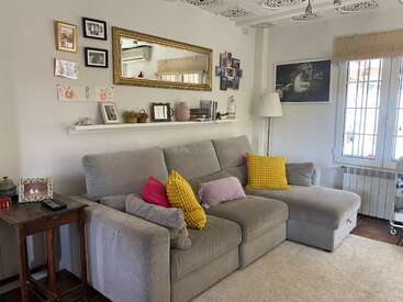 Un salón acogedor con un sofá modular gris, cojines de colores, cuadros, un espejo grande, fotos familiares, una mesita auxiliar de madera y luz natural.