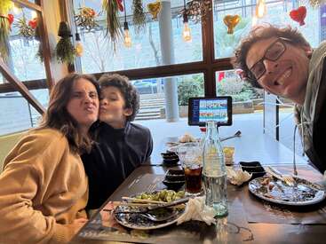 Una familia de tres personas está sentada en la mesa de un restaurante, con los platos ya vacíos, compartiendo un momento de diversión. Las luces decorativas y las plantas colgantes crean un ambiente acogedor y alegre.