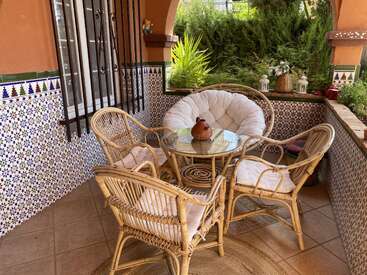Un acogedor patio exterior con cuatro sillas de mimbre alrededor de una mesa de cristal, paredes alicatadas con motivos coloridos, plantas verdes y farolillos decorativos, que crean un ambiente relajante.