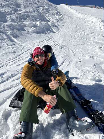Dos personas con ropa de esquí están sentadas juntas en la ladera nevada de una montaña. Una de ellas levanta el pulgar mientras sostiene una bebida. Ambas sonríen y disfrutan de un soleado día de invierno.