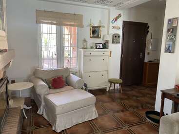 Un acogedor salón con un gran sillón y una otomana, muebles blancos, suelo de baldosas, adornos en las paredes, luz natural que entra por las ventanas y una puerta de entrada de madera oscura.