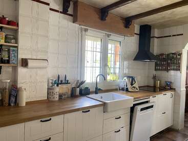 Cocina luminosa con armarios blancos, encimeras de madera, fregadero de estilo rústico, estante para especias, campana extractora negra, estantes abiertos, utensilios y una ventana que deja pasar la luz natural y ofrece vistas al jardín.