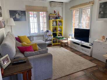 Un acogedor salón con sofás grises, cojines de colores, una estantería amarilla, un televisor sobre un mueble blanco, una mesa de juegos para niños, una alfombra, grandes ventanas y decoración en las paredes.