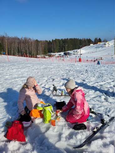 Two people in winter clothes sit on snow, enjoying a picnic with drinks and snacks. Ski equipment lies nearby. A snowy landscape and forest surround them.