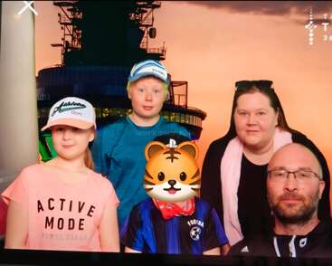 A family of five poses for a photo at sunset; three children, two adults. One child's face is covered with a tiger emoji. The background is industrial.