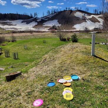 Colorful frisbees lie scattered on a grassy hill under a bright blue sky, with melting snow on a distant hill and trees in the background.
