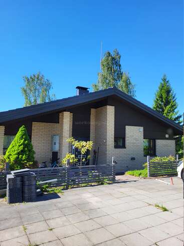 A modern single-story brick house with a dark roof, surrounded by greenery, trees, a small fence, and a paved driveway under a clear blue sky.