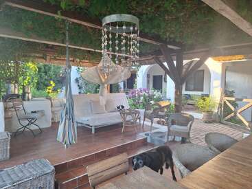 Un acogedor patio exterior con sillas de mimbre, un columpio, una lámpara de araña con cuentas de cristal, un sofá blanco, exuberante vegetación, plantas en macetas y un perro negro disfrutando del espacio.
