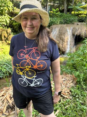 Une femme portant un chapeau de paille, un t-shirt de vélo "Budapest" et un short noir sourit dans un jardin verdoyant avec une cascade en arrière-plan.