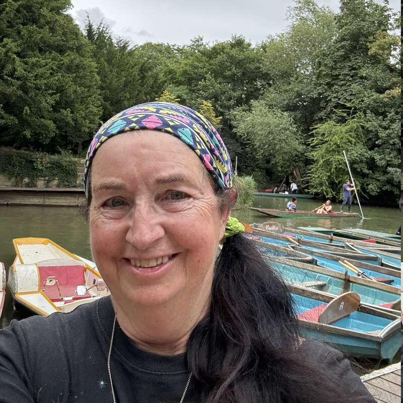 Une femme souriante portant un bandeau coloré prend un selfie au bord d'une rivière calme. Des bateaux à rames sont amarrés à proximité, avec des arbres verdoyants et des gens qui s'amusent à faire du bateau.