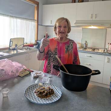 An elderly woman in a colorful top smiles and gives a thumbs up while cooking in a bright kitchen, proud of her dish on the counter.